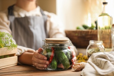 Woman holding glass jar of pickled cucumbers at wooden table, closeup