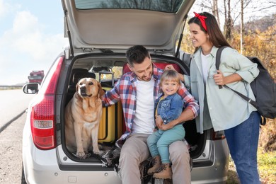 Photo of Parents, their daughter and dog sitting in car trunk outdoors. Family traveling with pet