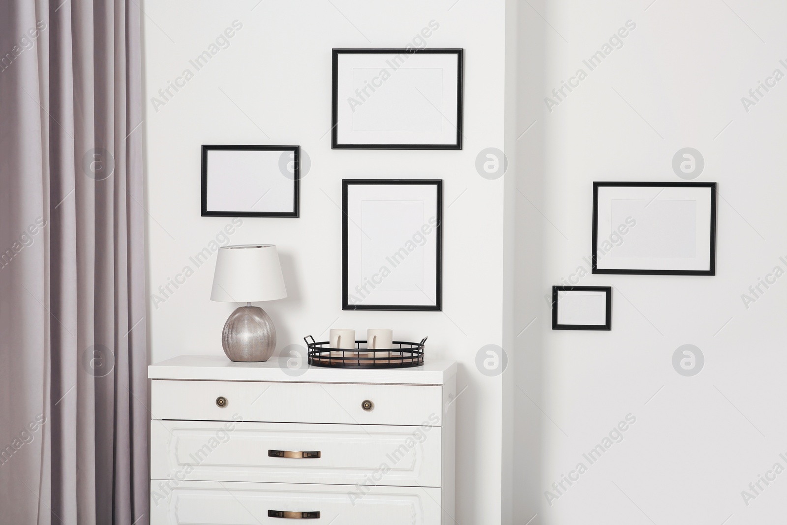 Photo of Empty frames hanging on white wall, wooden chest of drawers and lamp indoors