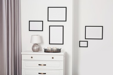 Photo of Empty frames hanging on white wall, wooden chest of drawers and lamp indoors