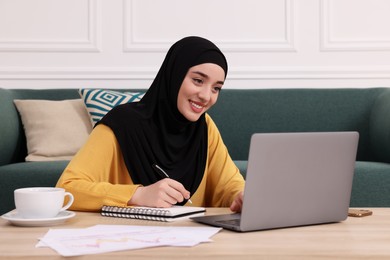 Photo of Muslim woman in hijab writing notes near laptop at wooden table indoors