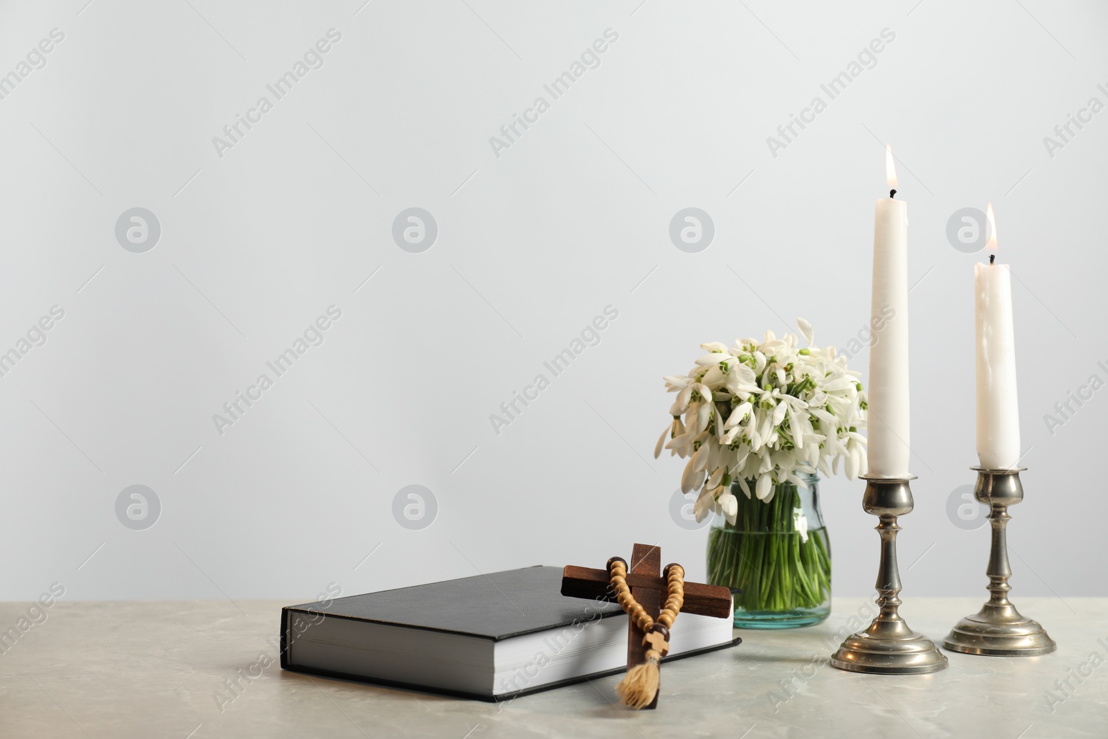 Photo of Church candles, wooden cross, rosary beads, Bible and flowers on marble table. Space for text