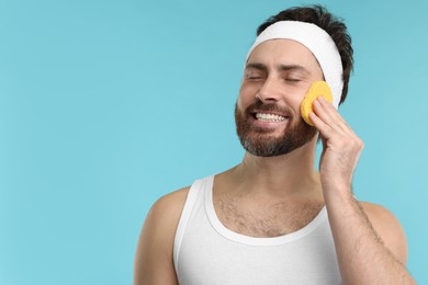 Photo of Man with headband washing his face using sponge on light blue background, space for text