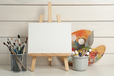 Photo of Easel with blank canvas, brushes, paints and palette on white wooden table