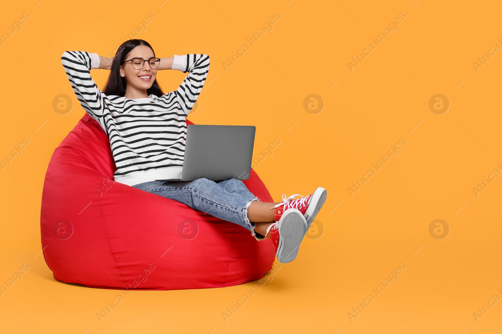 Photo of Happy woman with laptop sitting on beanbag chair against orange background