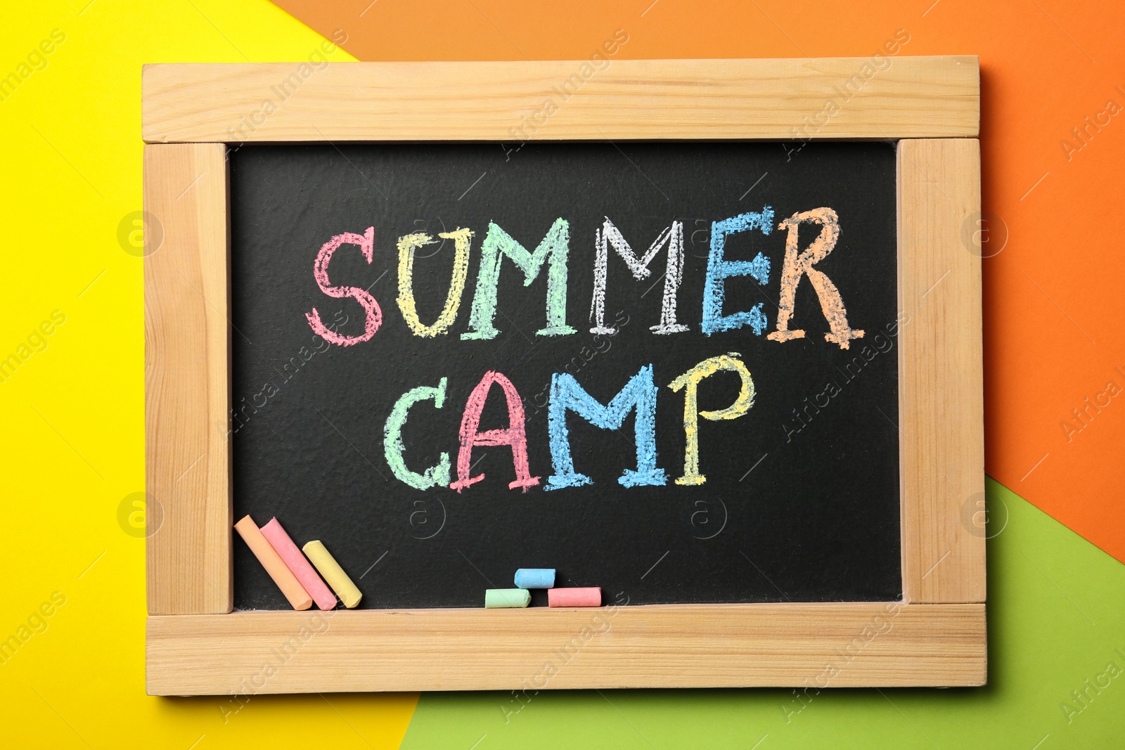 Photo of Small blackboard with text SUMMER CAMP and chalk sticks on color background, top view