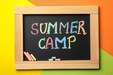 Small blackboard with text SUMMER CAMP and chalk sticks on color background, top view