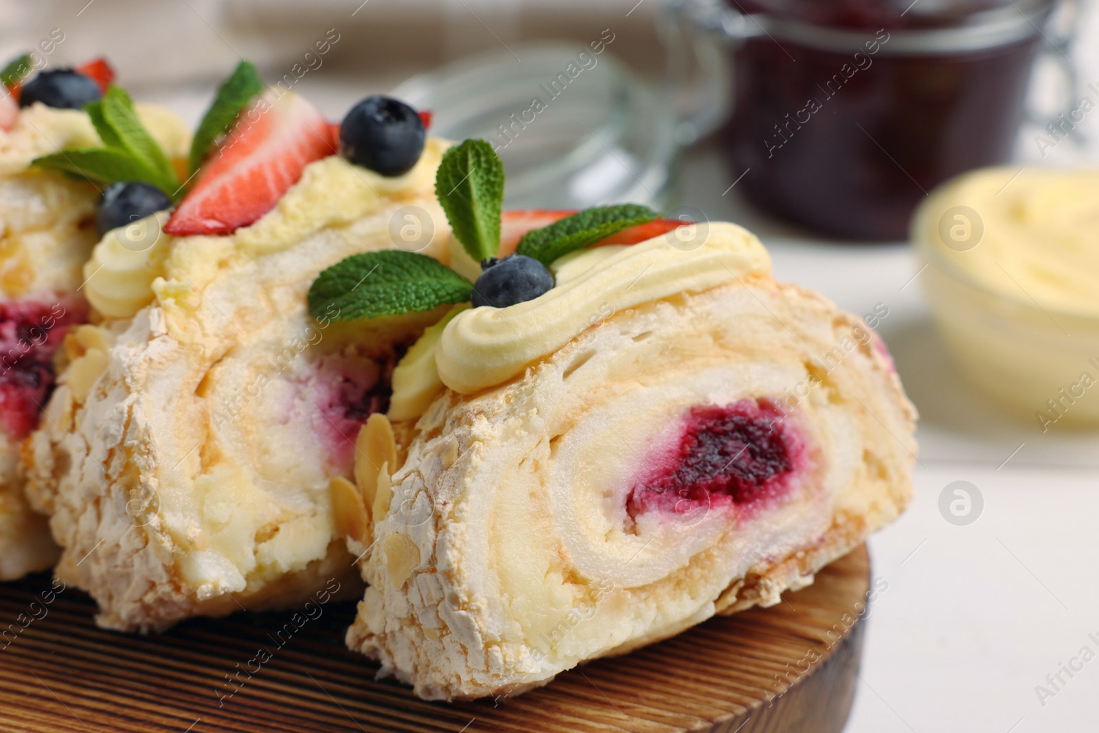 Photo of Pieces of tasty meringue roll with jam, cream, strawberry, blueberry and mint on table, closeup