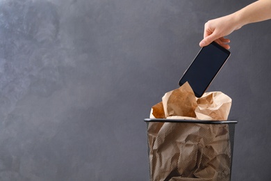 Closeup view of woman throwing smartphone into trash bin on grey background, space for text. Digital detox concept