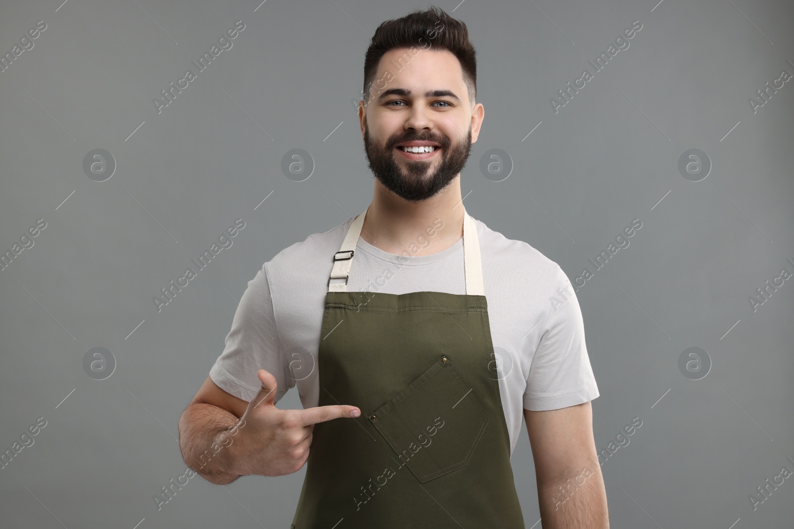 Photo of Smiling man pointing at kitchen apron on grey background. Mockup for design