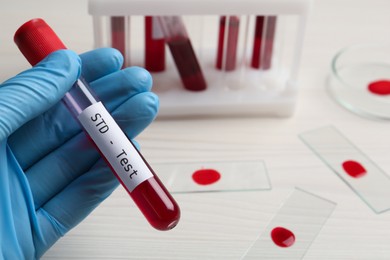 Scientist holding tube with blood sample and label STD Test at white table, closeup. Space for text