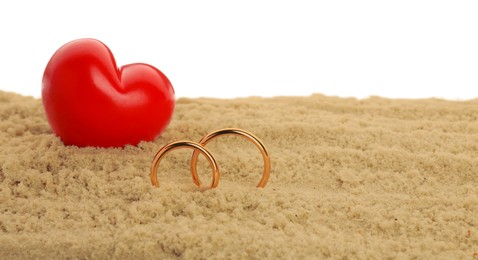 Photo of Honeymoon concept. Two golden rings, red wooden heart and sand isolated on white