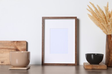 Photo of Empty photo frame, cups and vase with dry decorative spikes on wooden table. Mockup for design