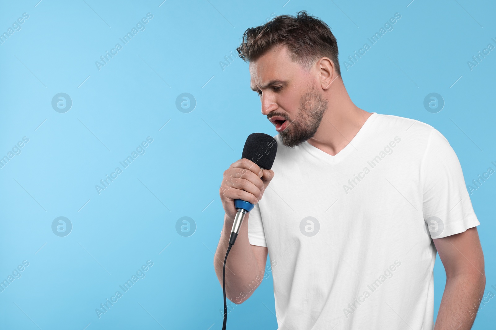 Photo of Handsome man with microphone singing on light blue background. Space for text
