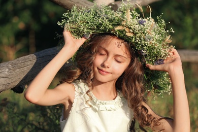 Photo of Cute little girl wearing wreath made of beautiful flowers near wooden fence outdoors on sunny day