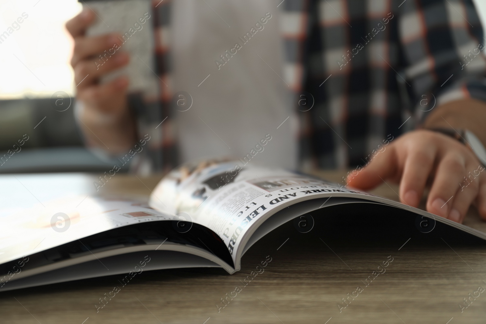 Photo of Man with cup of coffee reading fashion magazine at wooden table in room, closeup