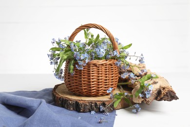 Beautiful forget-me-not flowers in wicker basket, blue cloth and piece of decorative wood on white table