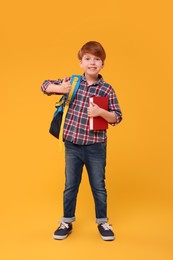 Photo of Smiling schoolboy with backpack and book showing thumb up on orange background