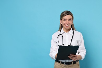 Portrait of happy doctor with stethoscope and clipboard on light blue background, space for text