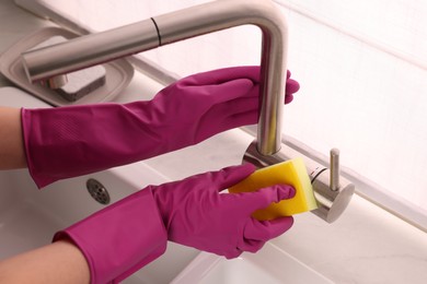 Photo of Woman in gloves cleaning faucet of kitchen sink with sponge, closeup