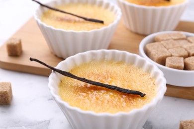 Photo of Delicious creme brulee in bowls, vanilla pods and sugar cubes on white marble table, closeup