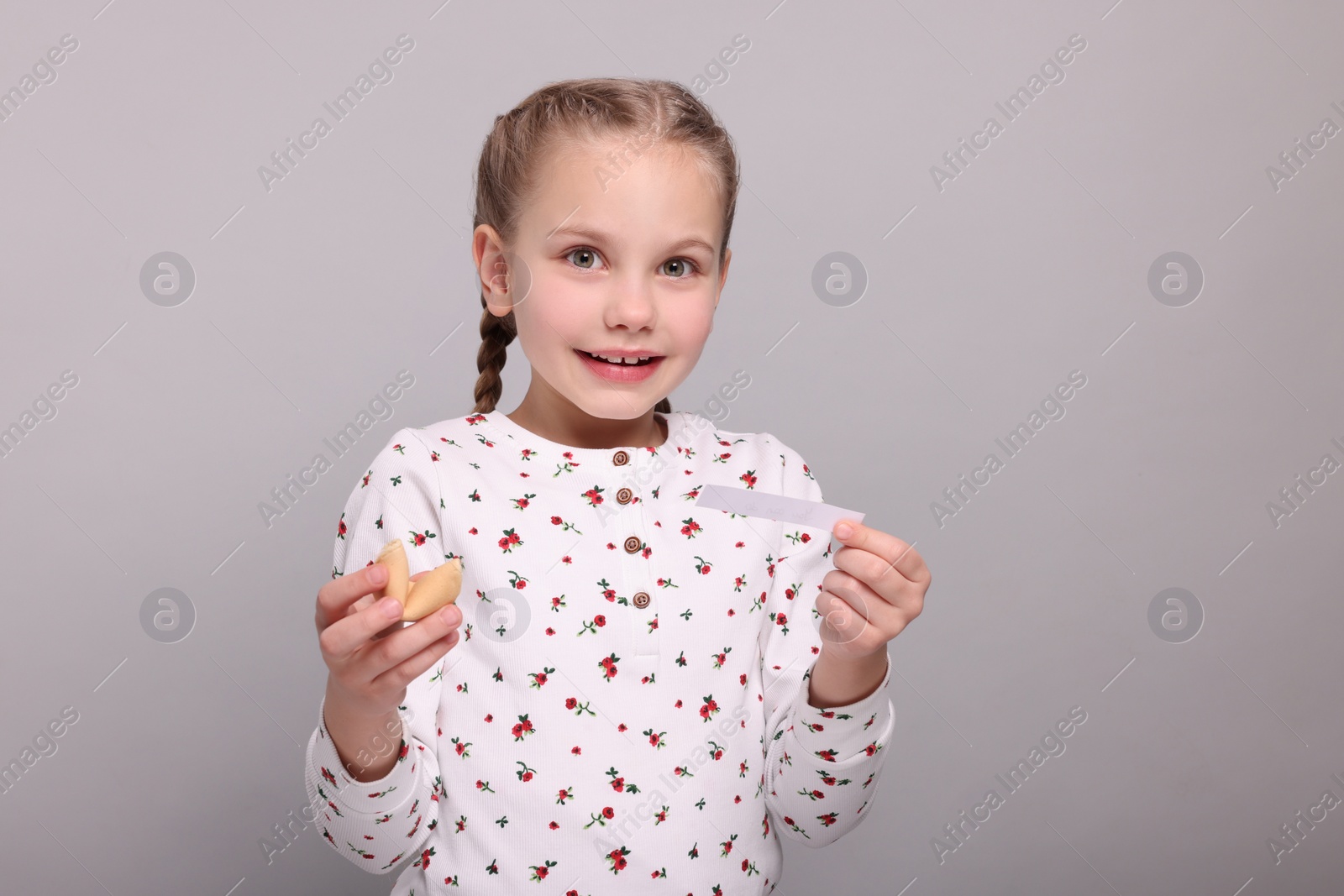 Photo of Cute girl holding tasty fortune cookie and reading prediction on light grey background