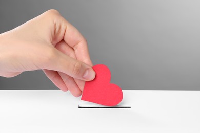 Photo of Woman putting red heart into slot of donation box against grey background, closeup