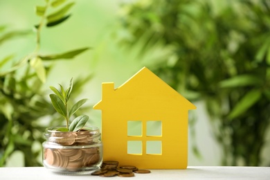 Photo of Model of house and jar with coins on table against blurred green background. Space for text