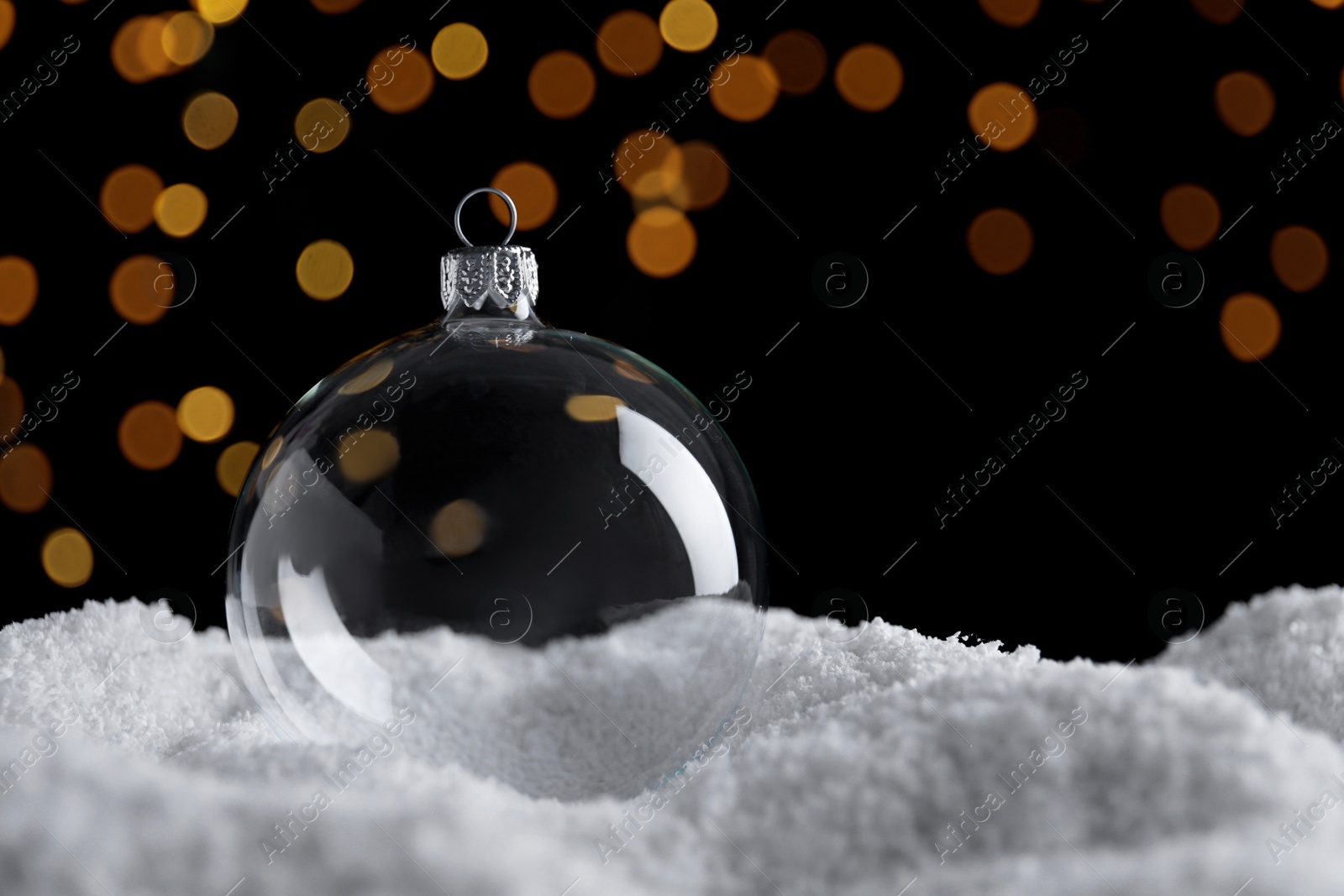 Photo of Transparent Christmas ball on snow against blurred fairy lights, space for text