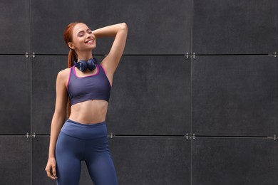 Beautiful woman in stylish gym clothes with headphones posing near dark grey wall on street, space for text