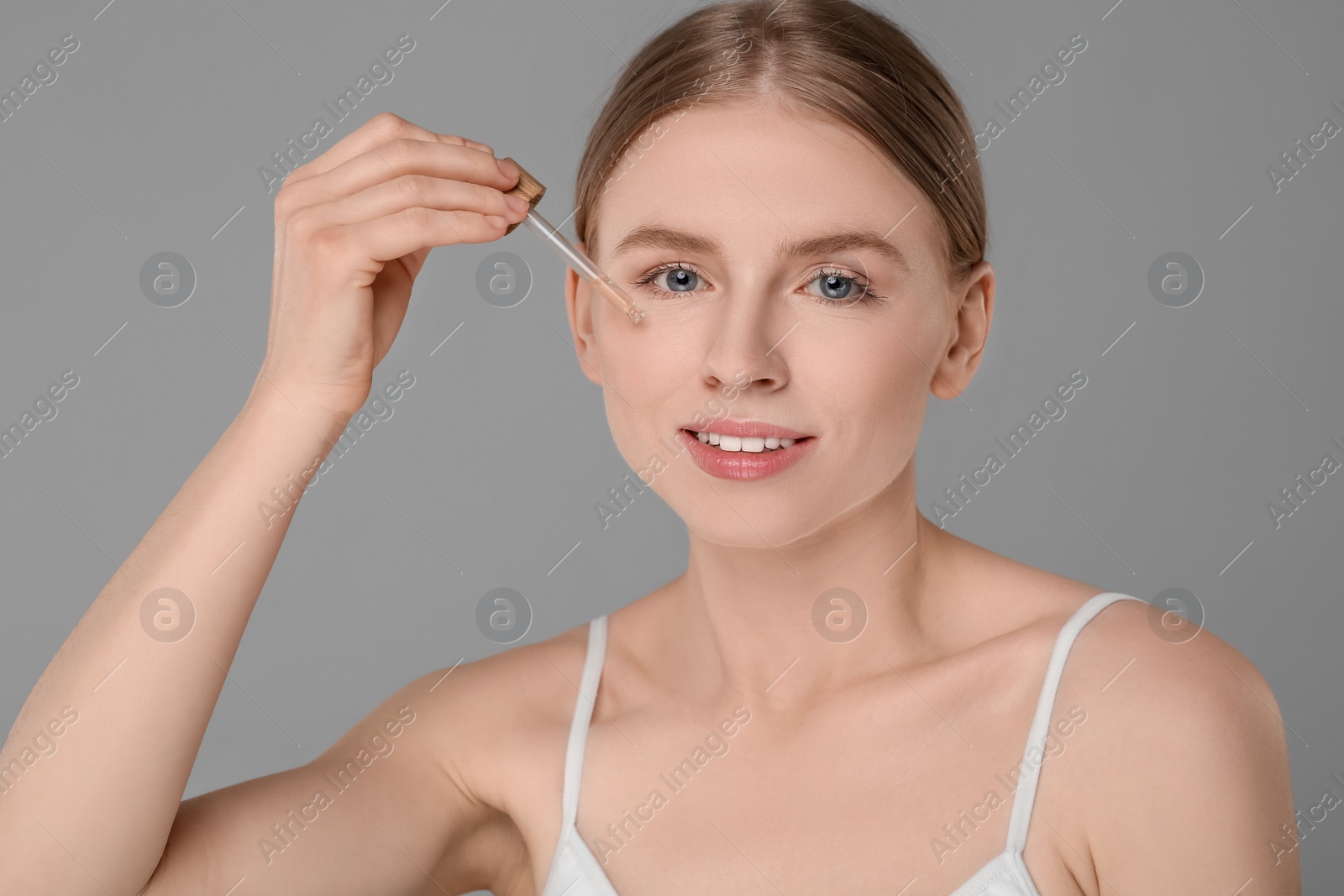 Photo of Beautiful young woman applying essential oil onto face on grey background