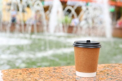 Photo of Cardboard cup with plastic lid on stone parapet near fountain outdoors, space for text. Coffee to go