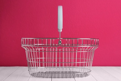 Photo of Empty metal shopping basket on white wooden table against crimson background