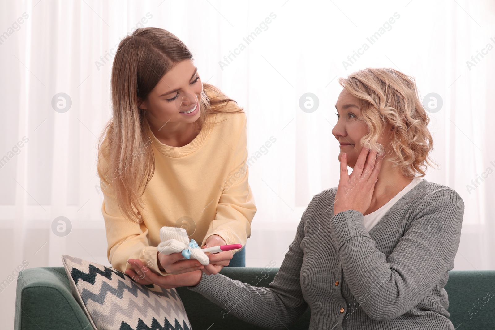 Photo of Young woman showing her mother pregnancy test and baby's socks at home. Grandparent reaction to future grandson