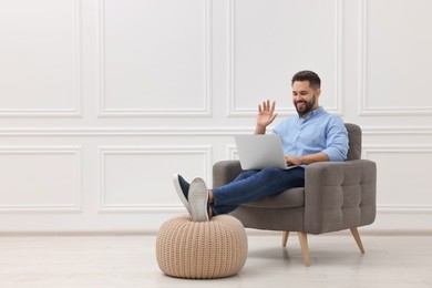 Photo of Handsome man having video chat via laptop while sitting in armchair near white wall indoors, space for text