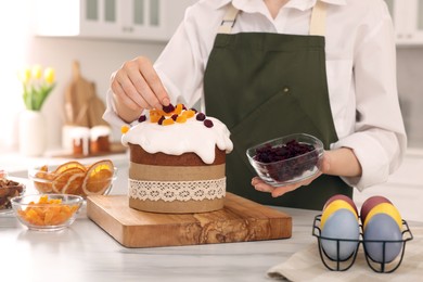 Woman decorating delicious Easter cake with dried cranberries at white marble table in kitchen, closeup