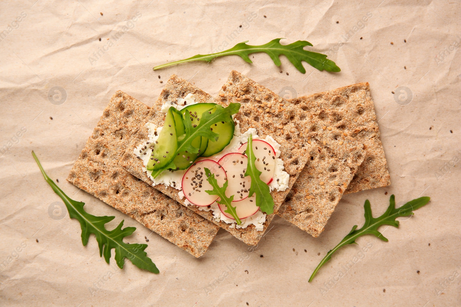 Photo of Tasty crispbreads with cream cheese, fresh cucumbers, radish and arugula on parchment paper, flat lay