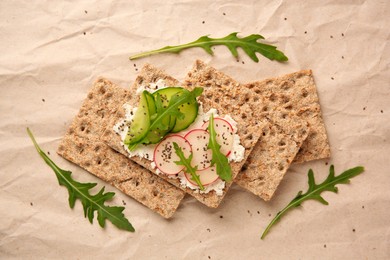 Tasty crispbreads with cream cheese, fresh cucumbers, radish and arugula on parchment paper, flat lay