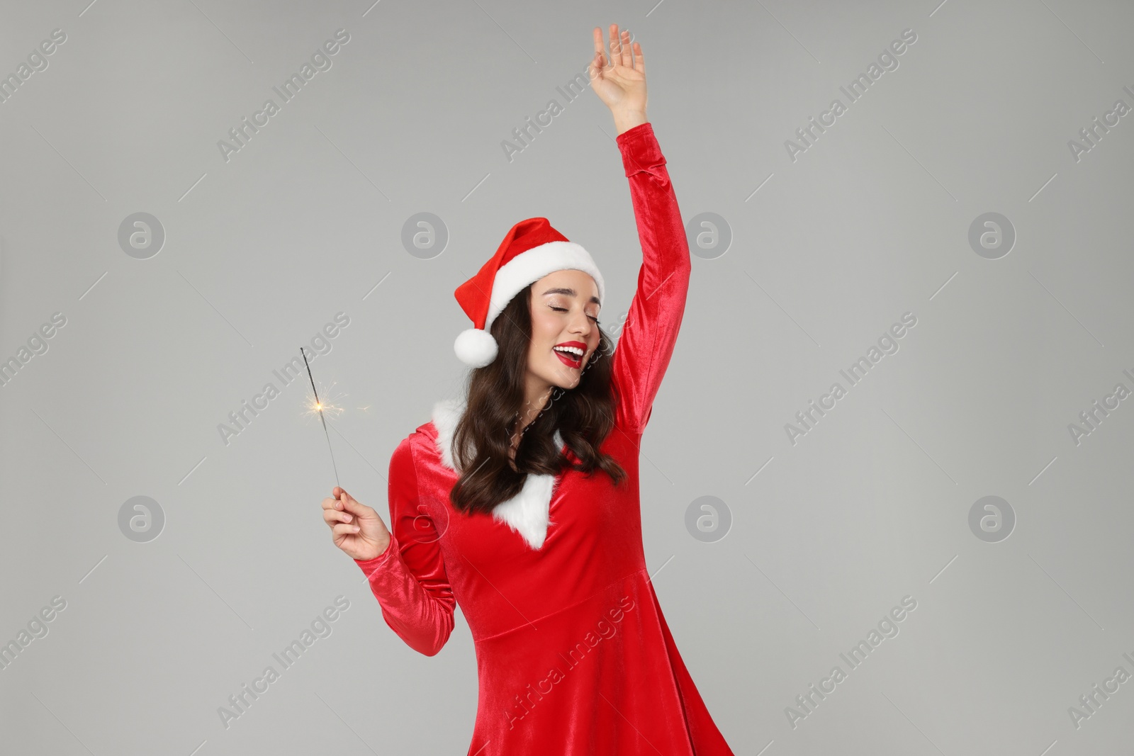 Photo of Christmas celebration. Beautiful young woman in red dress and Santa hat with sparkler on grey background