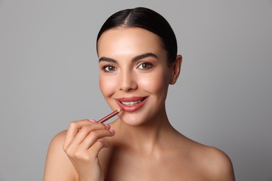 Photo of Pretty young woman applying beautiful nude lip pencil on grey background