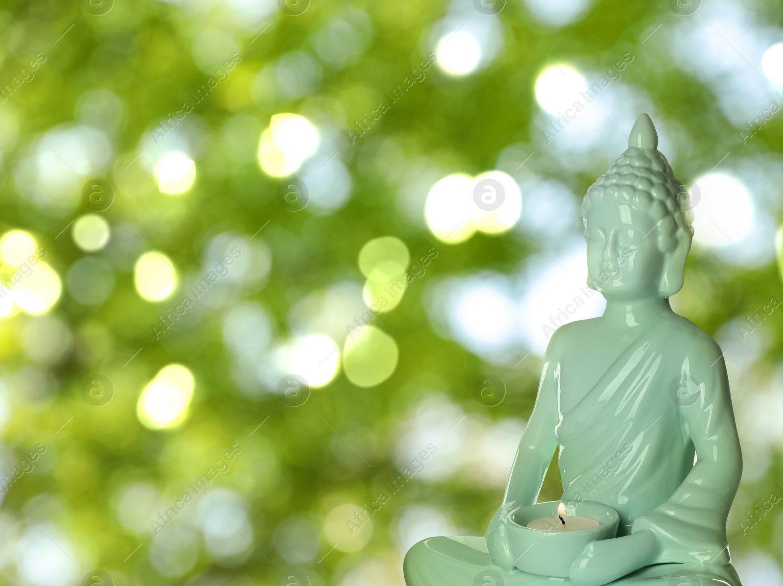 Image of Ceramic Buddha sculpture with burning candle outdoors. Space for text