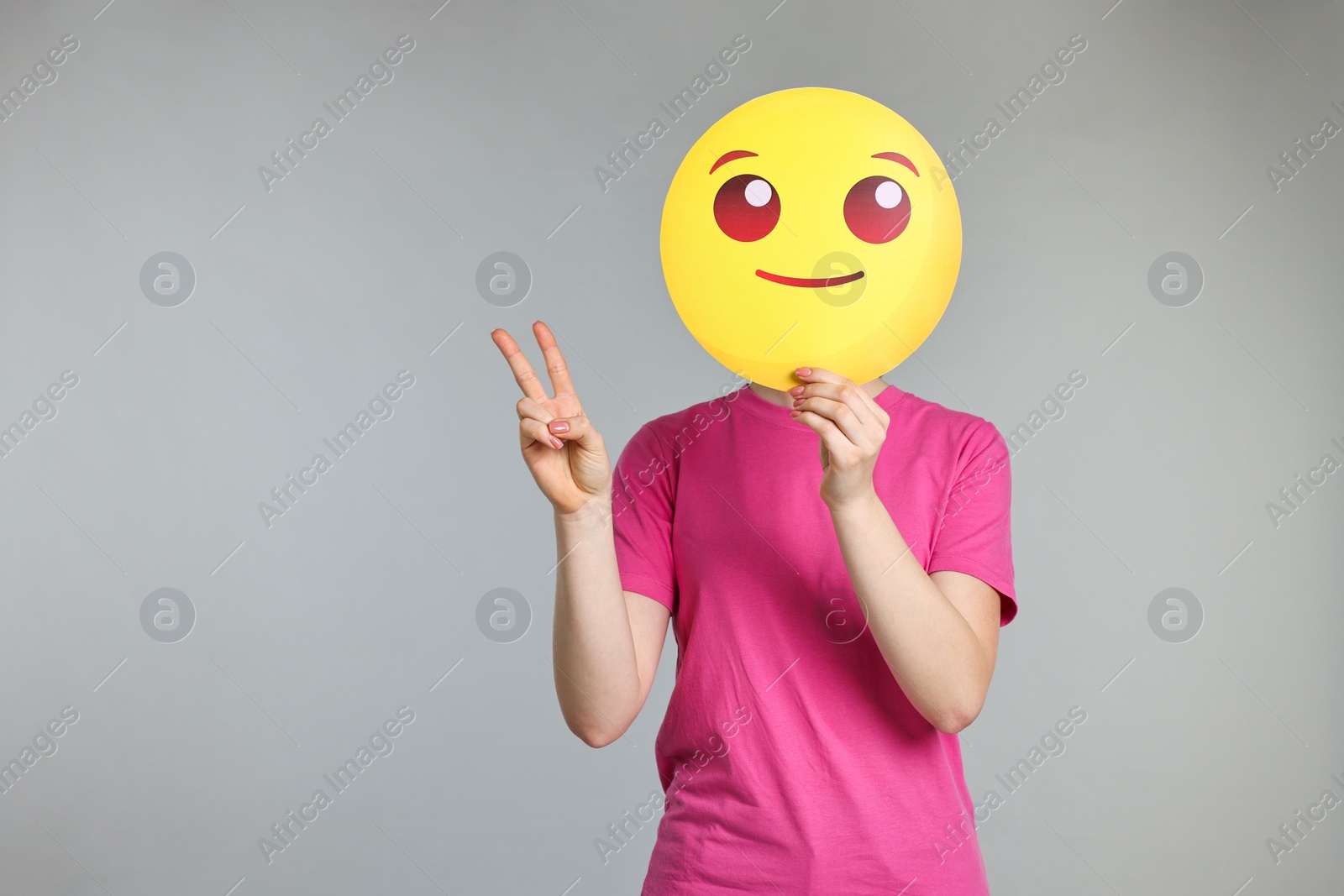 Photo of Woman covering face with smiling emoticon and showing peace sign on grey background. Space for text