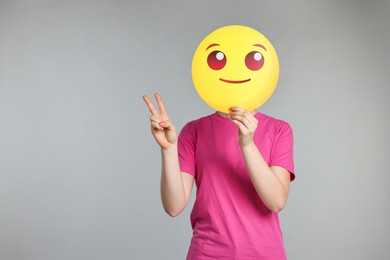 Photo of Woman covering face with smiling emoticon and showing peace sign on grey background. Space for text
