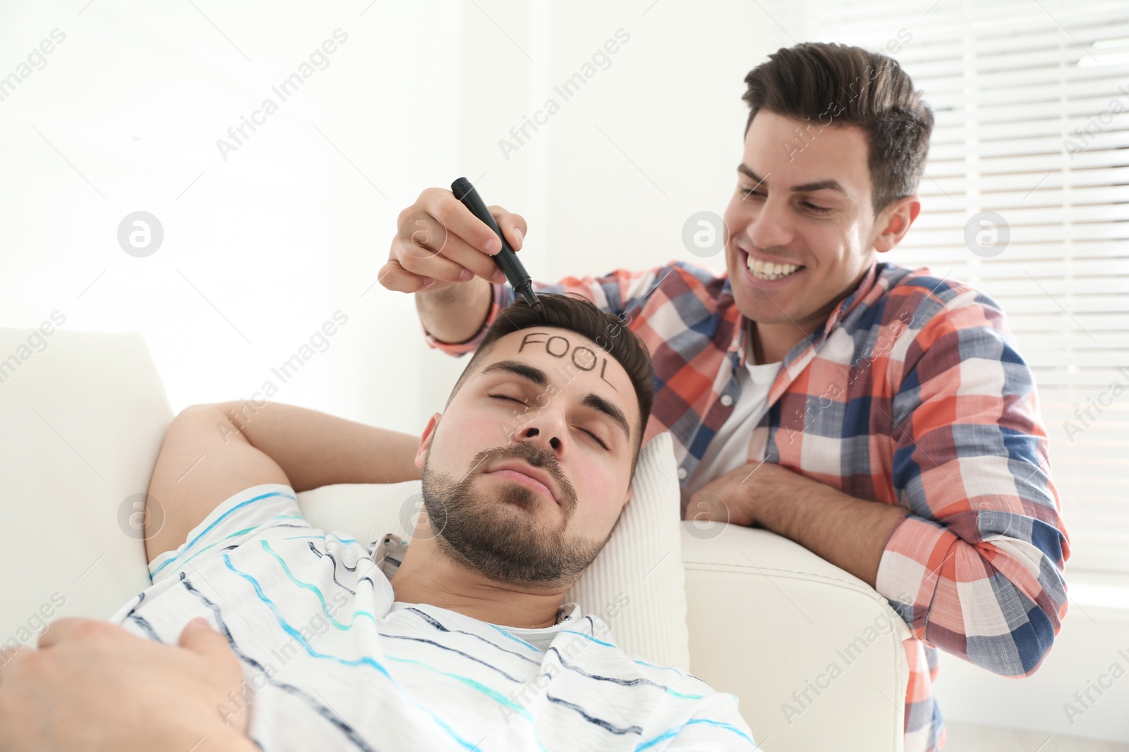 Photo of Man writing word FOOL on face of sleeping friend indoors. First April day