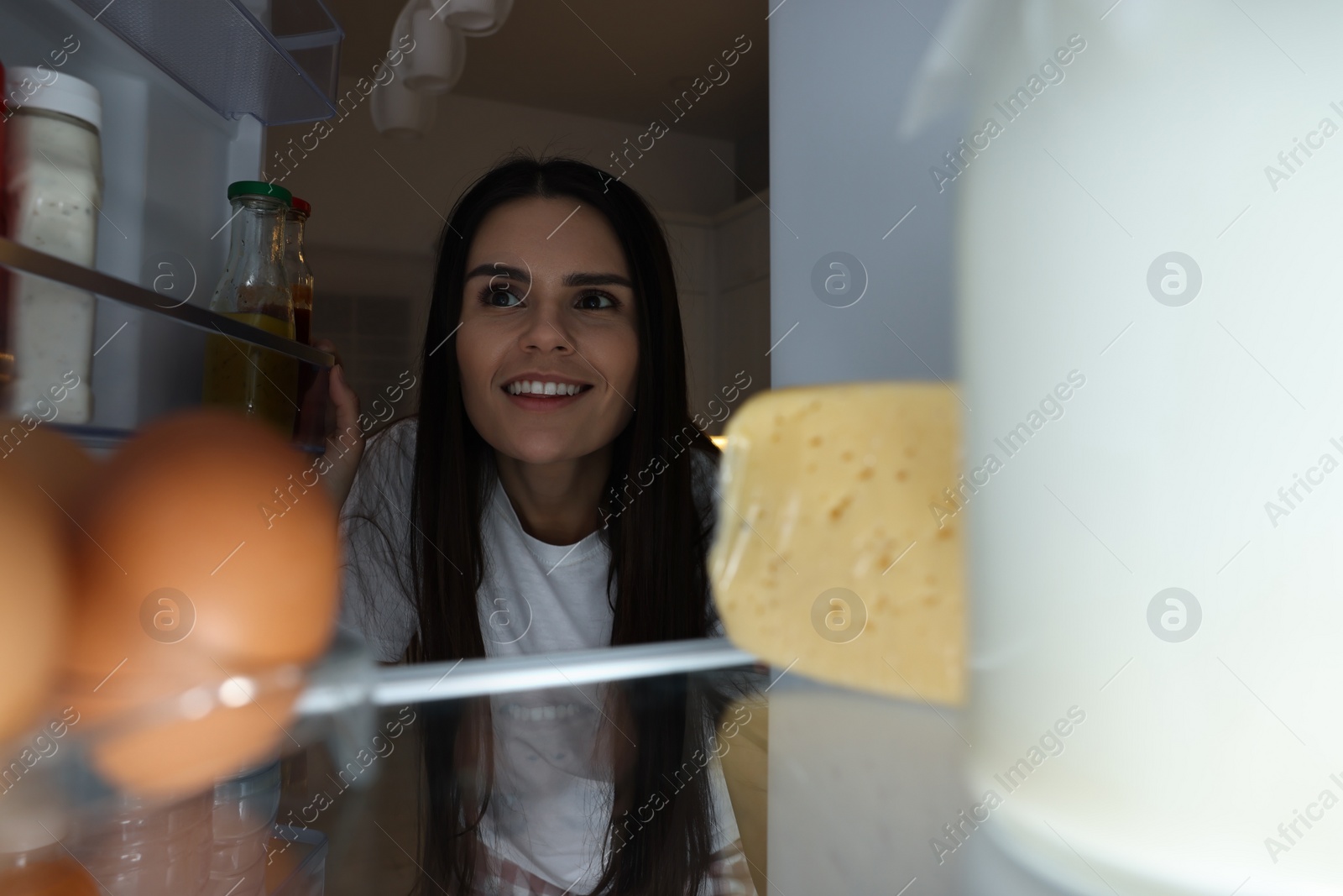 Photo of Young woman near modern refrigerator in kitchen at night, view from inside