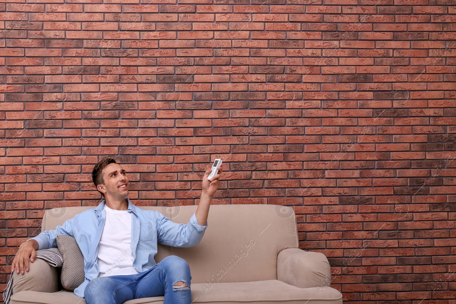 Photo of Young man operating air conditioner with remote control near brick wall at home