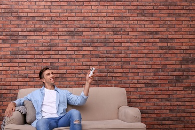 Young man operating air conditioner with remote control near brick wall at home