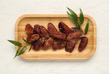 Plate with sweet dried dates and green leaves on white wooden table, top view