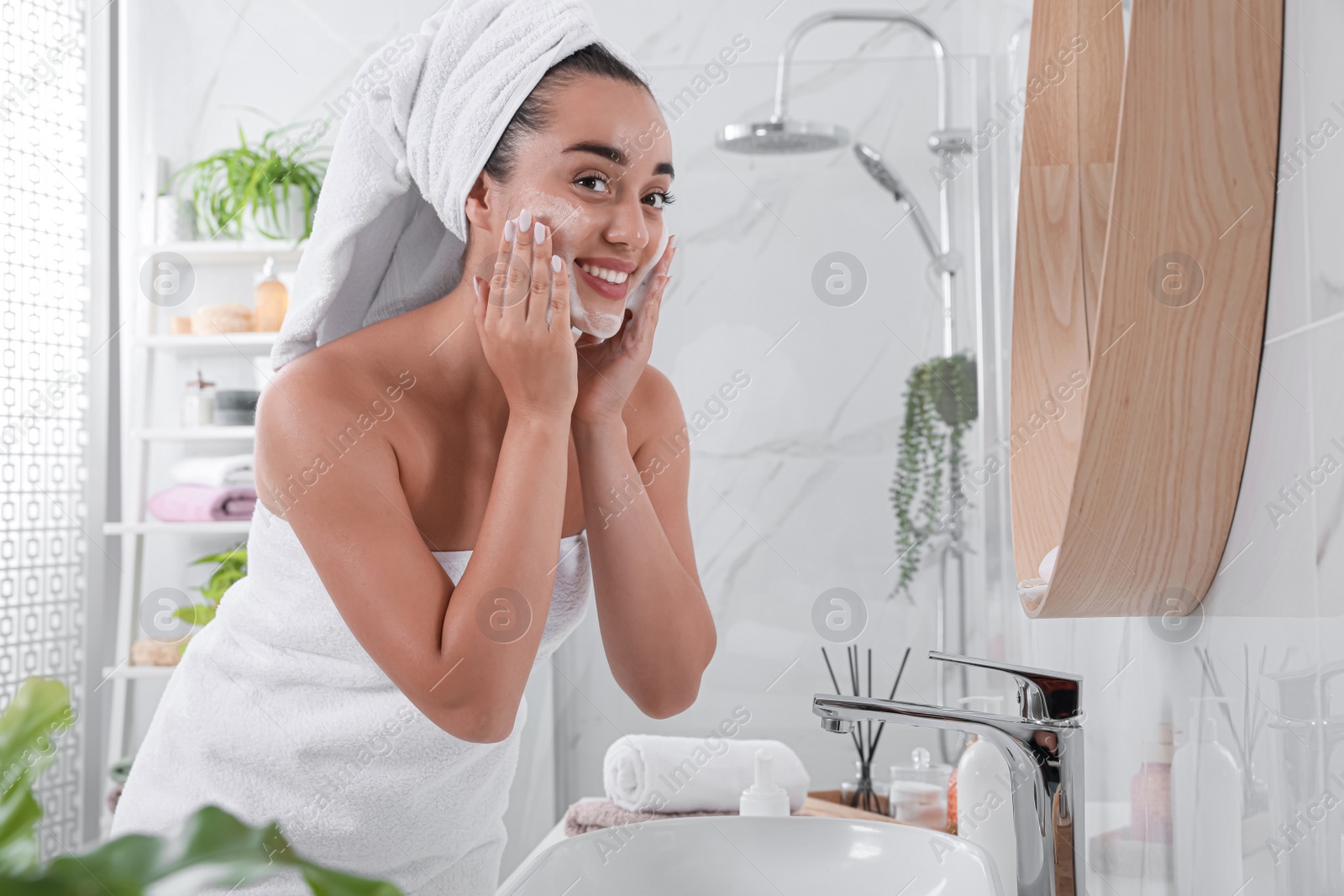 Photo of Beautiful young woman applying cleansing foam onto face near mirror in bathroom. Skin care cosmetic