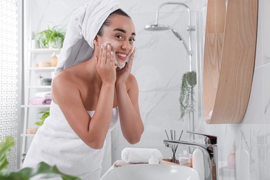 Beautiful young woman applying cleansing foam onto face near mirror in bathroom. Skin care cosmetic